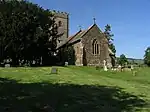 Stone building with square tower, partially obscured by trees. In the foreground are gravestones.