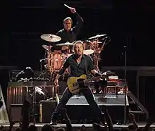 A man in dark clothing standing on a stage while holding a guitar in front of a crowd. Behind him is a man with his arm raised, holding a drum stick and sitting behind a drum set.