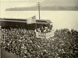 A dramatic political scene. Beside a river stands a podium, on which a flagpole flies a huge American flag. Beneath the flag stands a candidate in a dark suit addressing an impressive crowd that takes up most of the photograph. Not only the quayside, but a ferry beside it on the water are packed full of people listening intently.