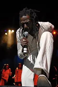 A man on a stage in white clothing, holding a microphone and bending over. In the background are women standing behind microphones.