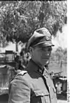 A man in semi profile standing in front of a tree and wearing a military uniform, peaked cap and a neck order in the shape of a cross. His cap has an emblem in shape of a human skull and crossed bones.