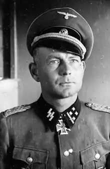 Black-and-white photo of a man wearing a peaked cap, military uniform with an Iron Cross displayed at his neck.