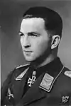 The head of a young man, shown in semi-profile. He wears a military uniform with an eagle above his right breast pocket, an iron cross is displayed at the front of his shirt collar. His hair is dark, short and combed to the back, his nose is long and straight, and his facial expression is determined; looking to the right of the camera.