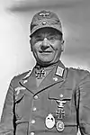 A man wearing a field cap and military uniform with various military decorations including an Iron Cross displayed at the front of his uniform collar.