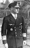 A man wearing a military uniform and peaked cap with a military order in shape of a cross displayed at the front of his uniform collar.