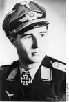 A man wearing a peaked cap, military uniform with an Iron Cross displayed at the front of his uniform collar.