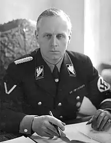 Portrait of a middle-aged man with short grey hair and a stern expression. He wears a dark military uniform, with a swastika on one arm. He is seated with his hands on a table with several papers on it, holding a pen.