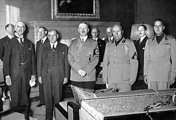 Interior photograph, showing in the foreground the five main signatories to the Munich Agreement, and in the background their various aides and assistants. Of the five, the two to the left are in black suits, the remainder in military uniform.