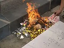 Pile of paper items on fire; a hand reaches to add money-like bills