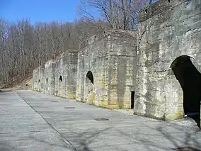 A row of six large stone structures with arched openings, leafless trees are in the background.