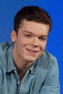 A 29-year-old man with long, brown hair, smiling at someone to the right of the camera.