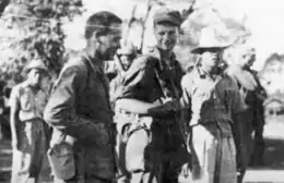 Black-and-white image of two men in military attire walking next to each other and smiling. Several other men can be seen in the background.