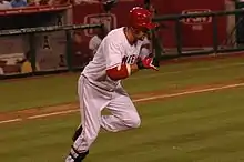 A dark-skinned man with "NGEL" visible in red text on a white jersey runs from home plate to first base. He has on red and black batting gloves, a red batting helmet, white pants and black shinguards.