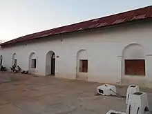 The white facade of a single-storey building, extending away from the viewer to the left. There is a single arched doorway, and a row of windows set into arched recesses. The roof is rusty corrugated metal. Four low white posts are linked by chains in the right foreground, one of them has been knocked over. Two ladies are visible in front of the left-most window. One sits on a dark plastic chair looking into a pram, the other stands behind the pram, also looking at it.