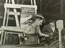 Black-and-white photograph of two soldiers working with a lathe. A poster behind them depicts a ship with hospital markings sinking by the bow and is captioned with "SAVE for the brave" and "Let us avenge the Nurses".