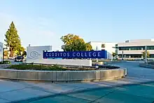 Sign for college with Cerritos College logo of a C within a C. Sign is in a pedestrian plaza with buildings in background.