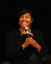 A woman with dark hair wearing a black outfit against a black background, she holds a microphone.