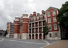 A four-story building of light grey stone and red brick with a complex facade including two central towers, and pillar and arch verandahs on the face of three of the four storeys