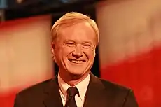 A man with yellow hair wearing a formal tie and suit, smiles with a series of red and white decorations behind him.