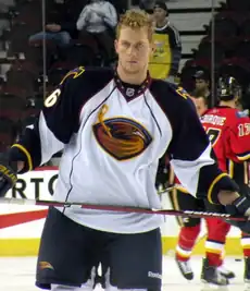 An ice hockey player stands directly upright holding an ice hockey stick horizontally across his stomach. He is wearing a no helmet and is wearing a black and white uniform with a large orange bird with an ice hockey stick on his chest.