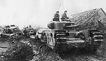 tanks moving along a muddy road beside a low building