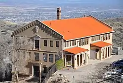 A large three-story building with many windows and an orange tile roof is perched on a hillside overlooking a wide valley far below.