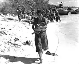 woman in dress in foreground walking toward viewer on a beach by the water's edge, soldiers and landing craft in background