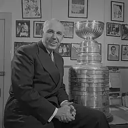 Photo of Campbell dressed in a suit and tie sitting on a desk with the Stanley Cup