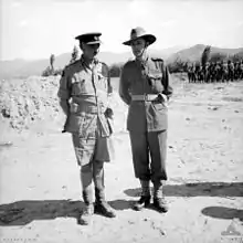 Two officers in tropical military uniforms standing on sandy ground, a larger body of troops is visible in the right background in front of stunted trees and more distant hills