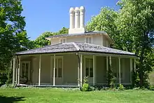 two-storey wooden building with wrap-around porch, surrounded by grassy area and trees