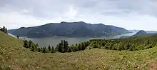 A wide river curves gently at the base of a mountain range. A meadow in the foreground gives way to an evergreen forest and then to the river. In the background, a layer of thin clouds veils a blue sky.