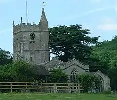 Gray stone building with arched windows. Square tower topped with spirelet, flagpole and weather vane. Foreground has small trees and bushes and a wooden rail fence.