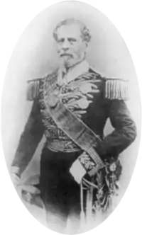 Tintype photograph depicting a full-length portrait of an older, bearded man wearing an elaborately embroidered military dress uniform with heavy epaulets, sash of office, sword, gloves and holding a bicorn hat in his right hand