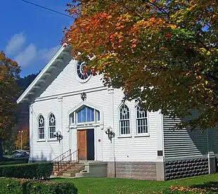 The front of a white building with a gabled roof. There are six-pointed Stars of David in the windows and a hedge around the walk leading to the stone steps to the front door, one of which is open. In the upper right the building is partially obscured by tree branches with orange, yellow and green leaves.