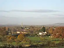 Roofs of houses showing amongst tree with prominent church tower. In the foreground are green fields with hills behind.