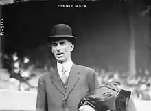 A man standing in a baseball stadium wearing a suit and bowler hat. He has a coat folded over his left arm.