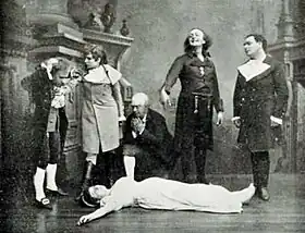 Photograph of white man with long hair in 19th century day clothes gesticulating manically over a female corpse, while two other men and a woman look on, horror-struck