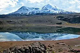 A snow-covered mountain with two hump-like summits rising above an unvegetated landscape with a lake