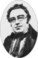 Round-faced white man, clean shaven with unruly dark hair and round spectacles