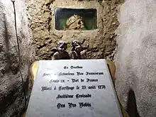 A human skull with a crown in a niche. Outside of the niche, two angel putti hold a marble plate inscribed 'Ex Ossibus / Sanctus Ludovicus Rex Francorum / Louis XI - Roi de France / Mort à Carthage le 25 août 1270 / Huitième Croisade / Ora Pro Nobis'.