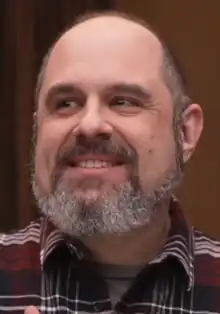 A 51 year-old man with a gray beard smiling to the left of the camera.