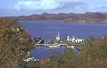 A small harbour surrounded by trees offers shelter to several yachts, fishing boats and a motor launch. The harbour is connected to the sea beyond by a lock gate. A small skerry lies in the water and brown rocky hills dominate the distance.