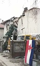 A blue-white-red Yugoslav flag, with the red Communist star in the middle, hangs on an iron fence outside the ruined shell of a house. A truck is partly visible parked in the driveway next to the building.