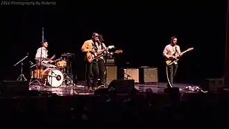 Wide shot of a four-piece band performing on stage. Left to right: drums; Harding, guitar and microphone; bass; and guitar