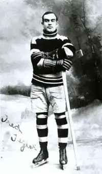 A young man poses for a photo, wearing a sweater, skates, and holding an ice hockey stick on his left side