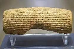 Rear view of a barrel-shaped clay cylinder resting on a stand. The cylinder is covered with lines of cuneiform text