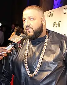 A man with short brown-blond hair and a dark beard speaks into a wireless microphone. He is wearing a shiny black shirt and has a dangling gold chain around his neck.