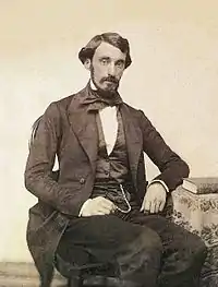 Daguerrotype portrait of a man seated next to a table and wearing black suit, vest and tie.