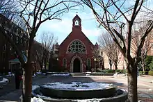 A frontal view of the red, brick edifice of the chapel. In front is a brick quadrangle with some snow on the ground, all centered between two trees in the foreground.