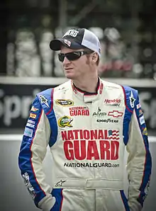 A man in his thirties wearing a white jacket with sponsors logos and a black and white baseball cap. He has both his hands in his pockets.